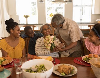 Front view of African American senior man giving a beautiful flower bouquet to his wife surrounded by her daughter and her grandchildren while having meal on dining table at home