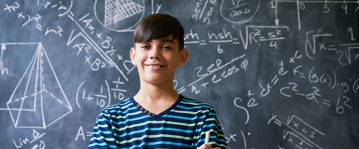 Concept on blackboard at school. Young people, student and pupil in classroom. Smart hispanic boy writing math formula on board during lesson. Portrait of male child smiling, looking at camera