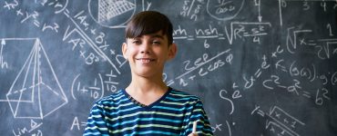 Concept on blackboard at school. Young people, student and pupil in classroom. Smart hispanic boy writing math formula on board during lesson. Portrait of male child smiling, looking at camera