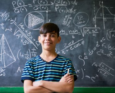 Concept on blackboard at school. Young people, student and pupil in classroom. Smart hispanic boy writing math formula on board during lesson. Portrait of male child smiling, looking at camera