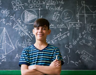 Concept on blackboard at school. Young people, student and pupil in classroom. Smart hispanic boy writing math formula on board during lesson. Portrait of male child smiling, looking at camera