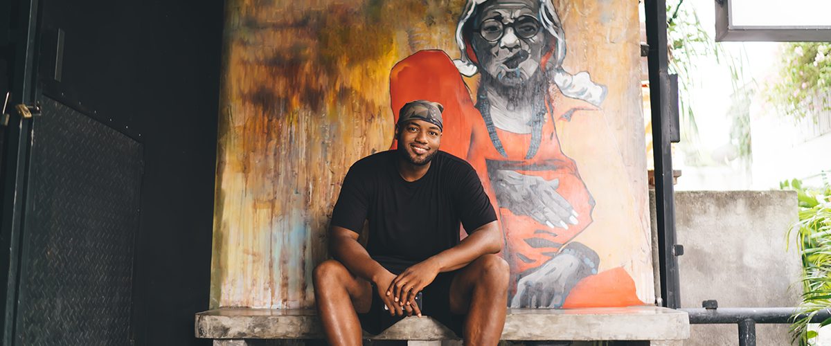 Portrait of happy African American hipster guy dressed in casual clothes smiling at camera while resting at sidewalk bench with creative graffiti illustration on background wall, hip hop street art