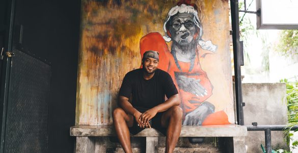 Portrait of happy African American hipster guy dressed in casual clothes smiling at camera while resting at sidewalk bench with creative graffiti illustration on background wall, hip hop street art