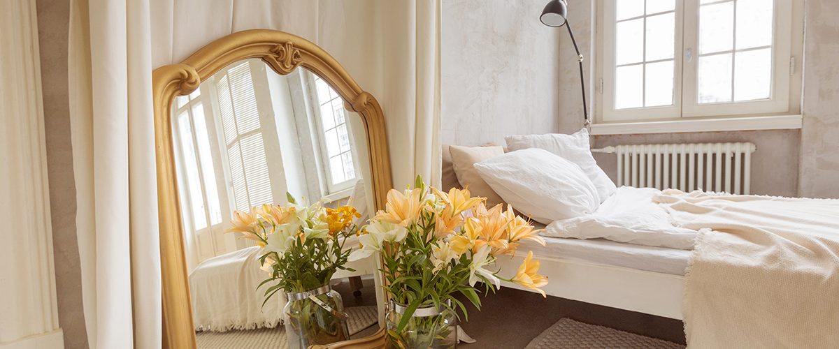 Bouquet of lilies flowers and mirror placed near curtain and comfortable bed in elegant bedroom at home