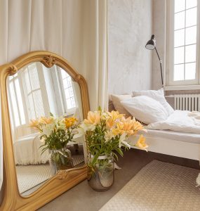 Bouquet of lilies flowers and mirror placed near curtain and comfortable bed in elegant bedroom at home