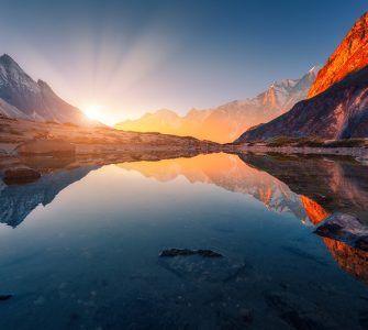 Beautiful landscape with high mountains with illuminated peaks, stones in mountain lake, reflection, blue sky and yellow sunlight in sunrise. Nepal. Amazing scene with Himalayan mountains. Himalayas