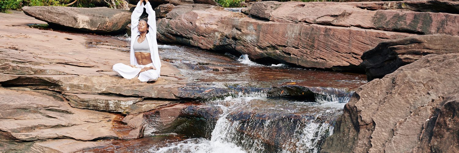 woman in white practicing awareness yoga next to waterfall cascading from red rocks