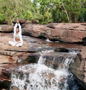 woman in white practicing awareness yoga next to waterfall cascading from red rocks