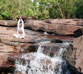 woman in white practicing awareness yoga next to waterfall cascading from red rocks