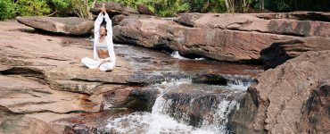 woman in white practicing awareness yoga next to waterfall cascading from red rocks