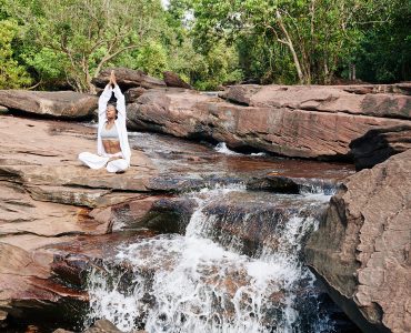 woman in white practicing awareness yoga next to waterfall cascading from red rocks