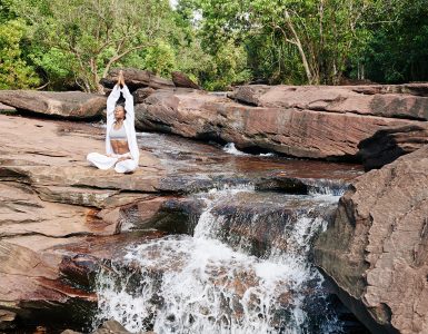 woman in white practicing awareness yoga next to waterfall cascading from red rocks