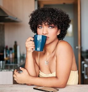 Transgender woman looking away while drinking a cup of coffee sitting in the kitchen at home.