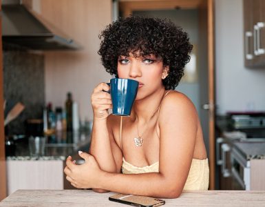 Transgender woman looking away while drinking a cup of coffee sitting in the kitchen at home.