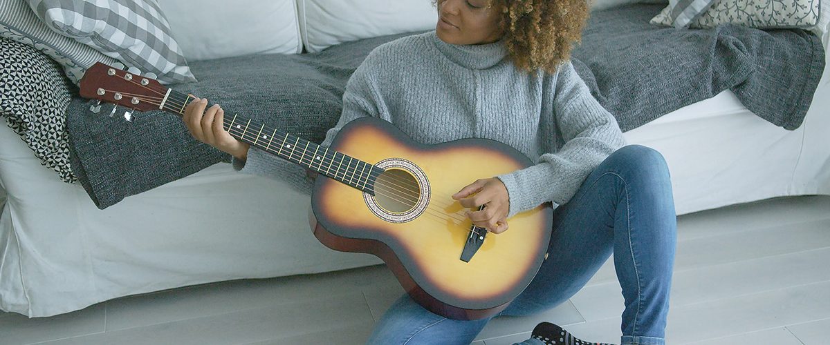 Charming young model in cozy sweater sitting on floor of living room and playing guitar alone looking inspired.