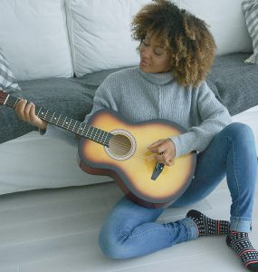 Charming young model in cozy sweater sitting on floor of living room and playing guitar alone looking inspired.