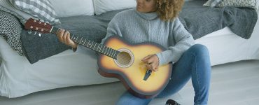 Charming young model in cozy sweater sitting on floor of living room and playing guitar alone looking inspired.